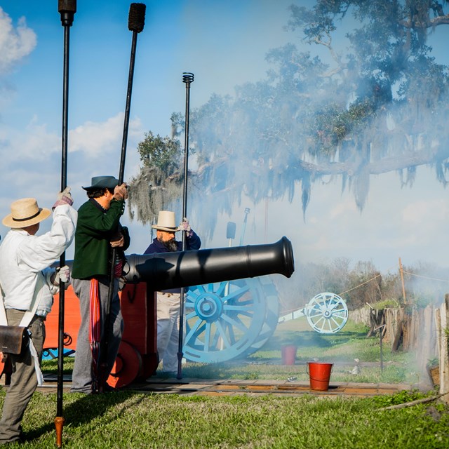 a group of reenactors dressed in period dress stand next to a cannon with smoke filling the air