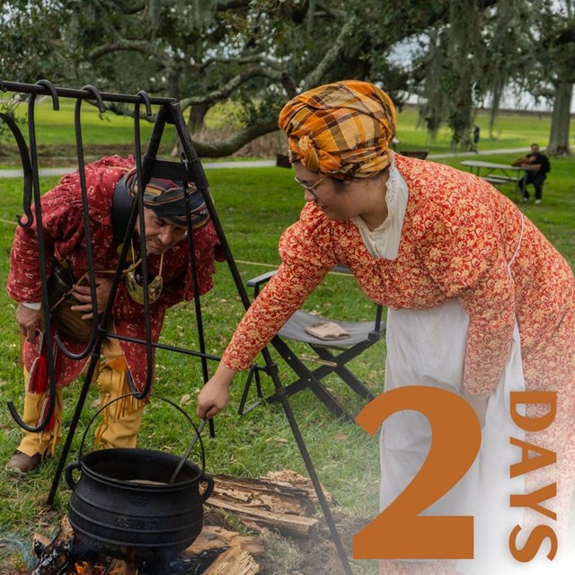Woman in reenactment clothing holds a spoon in a black pot over wood. A man peers into the pot.