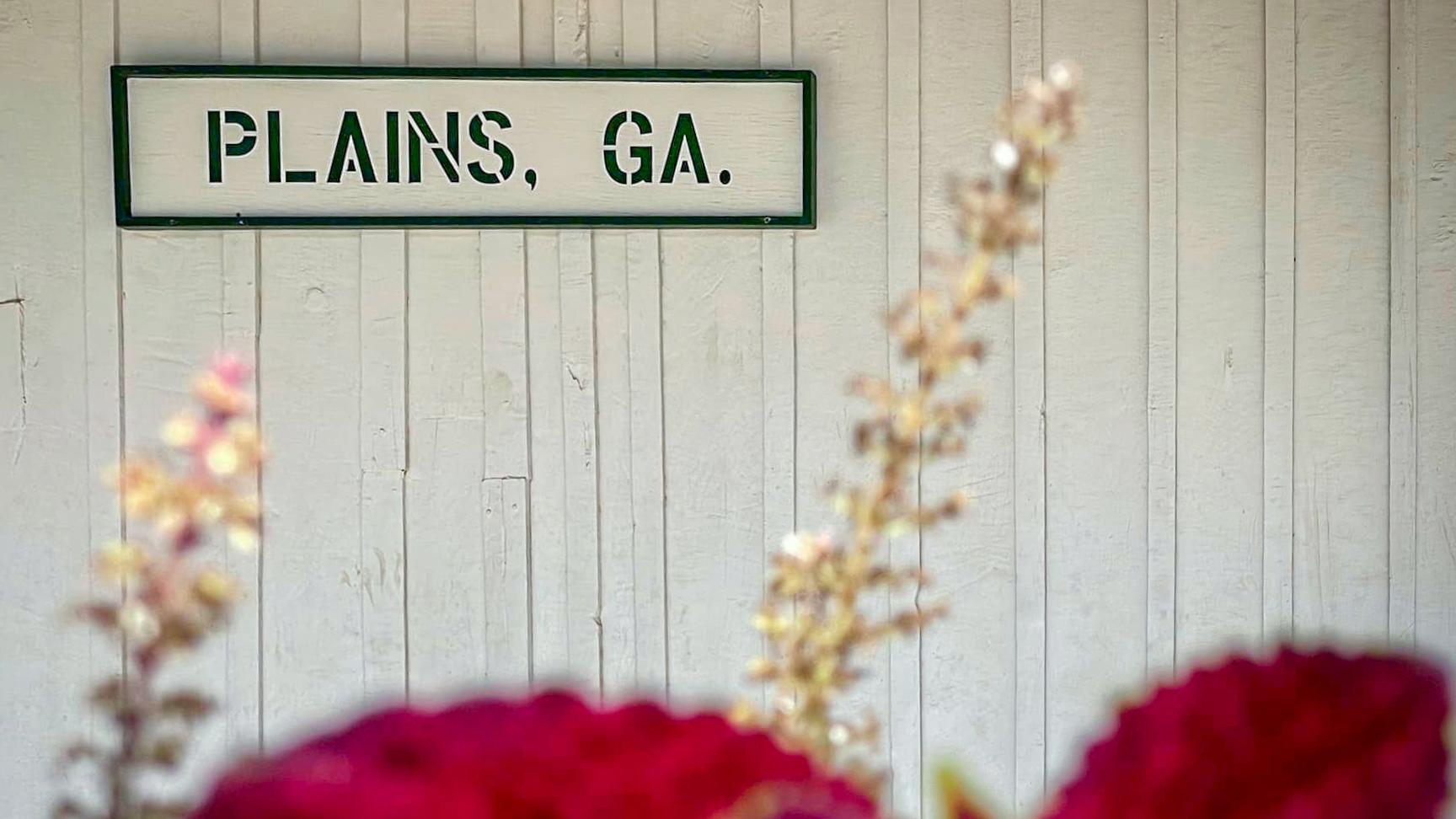Plains, GA sign on wall at Plains Depot with flowers. 