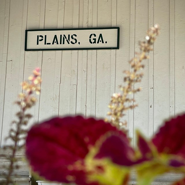 Plains, GA sign on wall at Plains Depot with flowers. 
