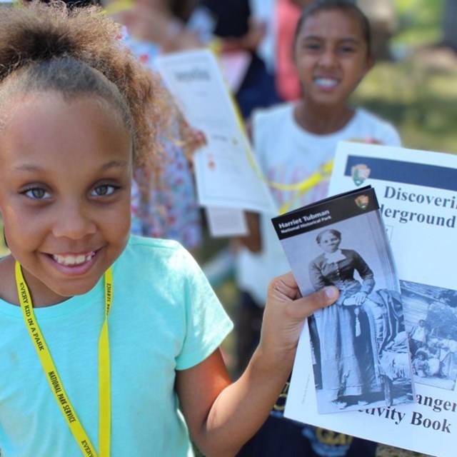 A young, smiling girl holds a Junior Ranger booklet titled Discovering the Underground Railroad.