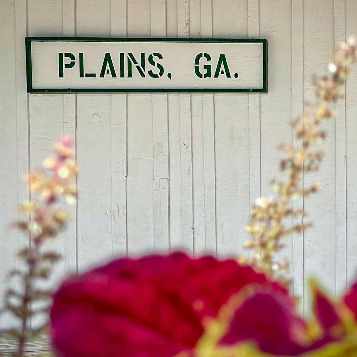 Plains, GA sign on wall at Plains Depot with flowers. 
