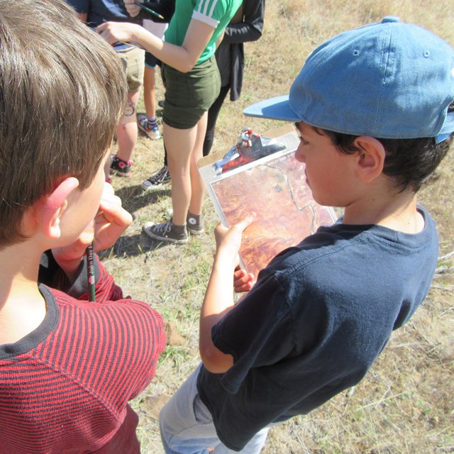 several students hold clipboards while listening to a park ranger outside