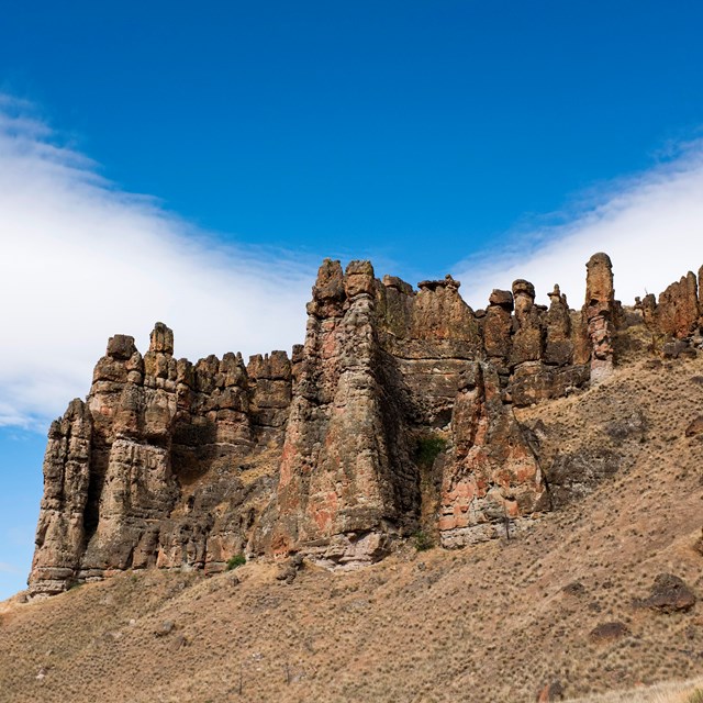 Brown mudflow cliff formations on top of a grassy slope with cloudy blue skies above.