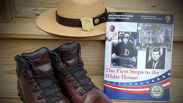 A pair of hiking boots, a park ranger flat hat, and the JFK NHS Junior Ranger booklet are displayed 