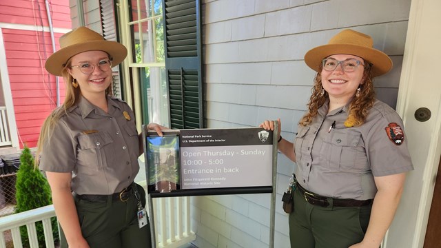 Photo of two Park Rangers standing in front of an open sign with hours in front of 83 Beals Street.
