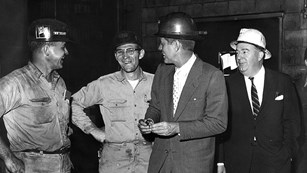 JFK, wearing a suit and hardhat stands with miners wearing hard hats