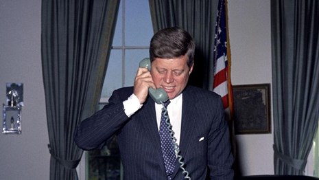 President Kennedy talks on the phone in the oval office, standing behind his desk. 