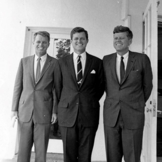 3 men in suits, Robert, Ted, and John F. Kennedy, stand outside the oval office. 
