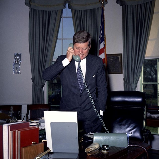President Kennedy talks on the phone in the oval office, standing behind his desk. 