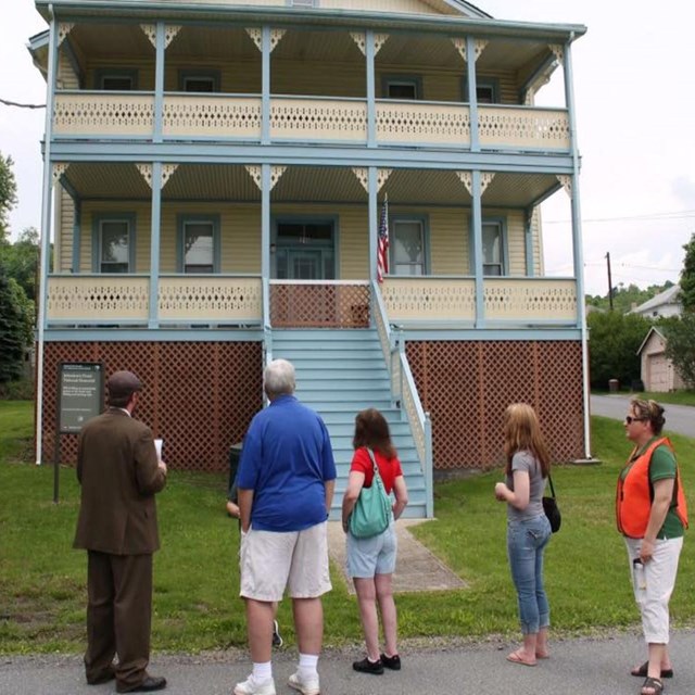 A park ranger in period clothing talks to a group in front of a cottage.