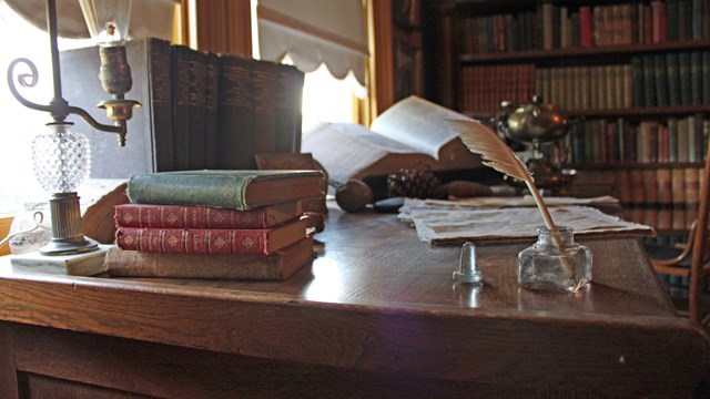 A desk is seen with piles of books and various items. Light shines through a window. 