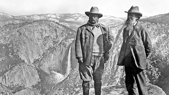 Two men in suits stand on a mountain. One beard, one mustache and glasses. 
