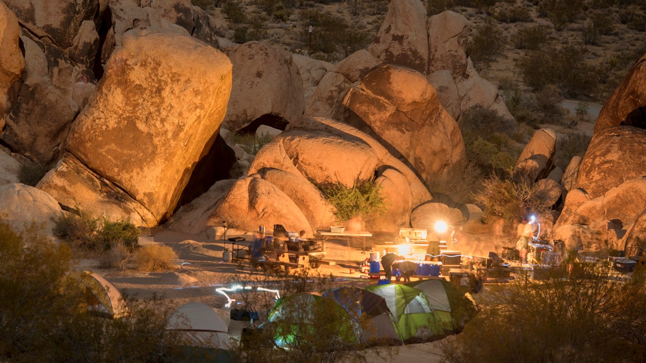 Color photo of a campsite at dark with a fire lit and headlamps leaving light trails.