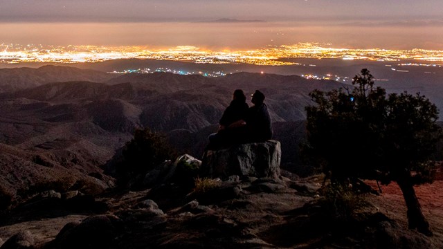 a couple sitting at a viewpoint at night, looking toward the lights of the Coachella Valley