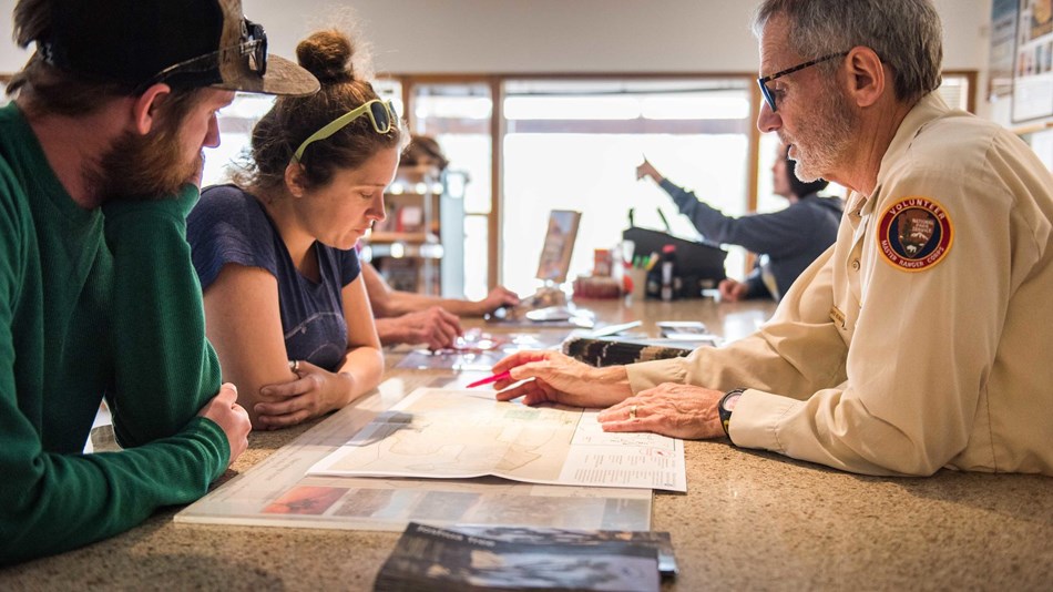 A park volunteer and two visitors look at a map of the park in a visitor center. 