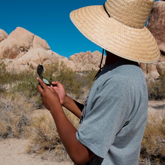 Young man wearing a large sun hat faces away from the camera holding a compass.