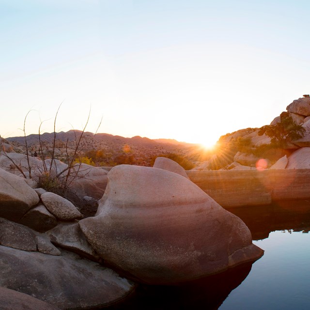 Color photo of water behind the man-made Barker Dam with the sun low on the horizon at sunset.