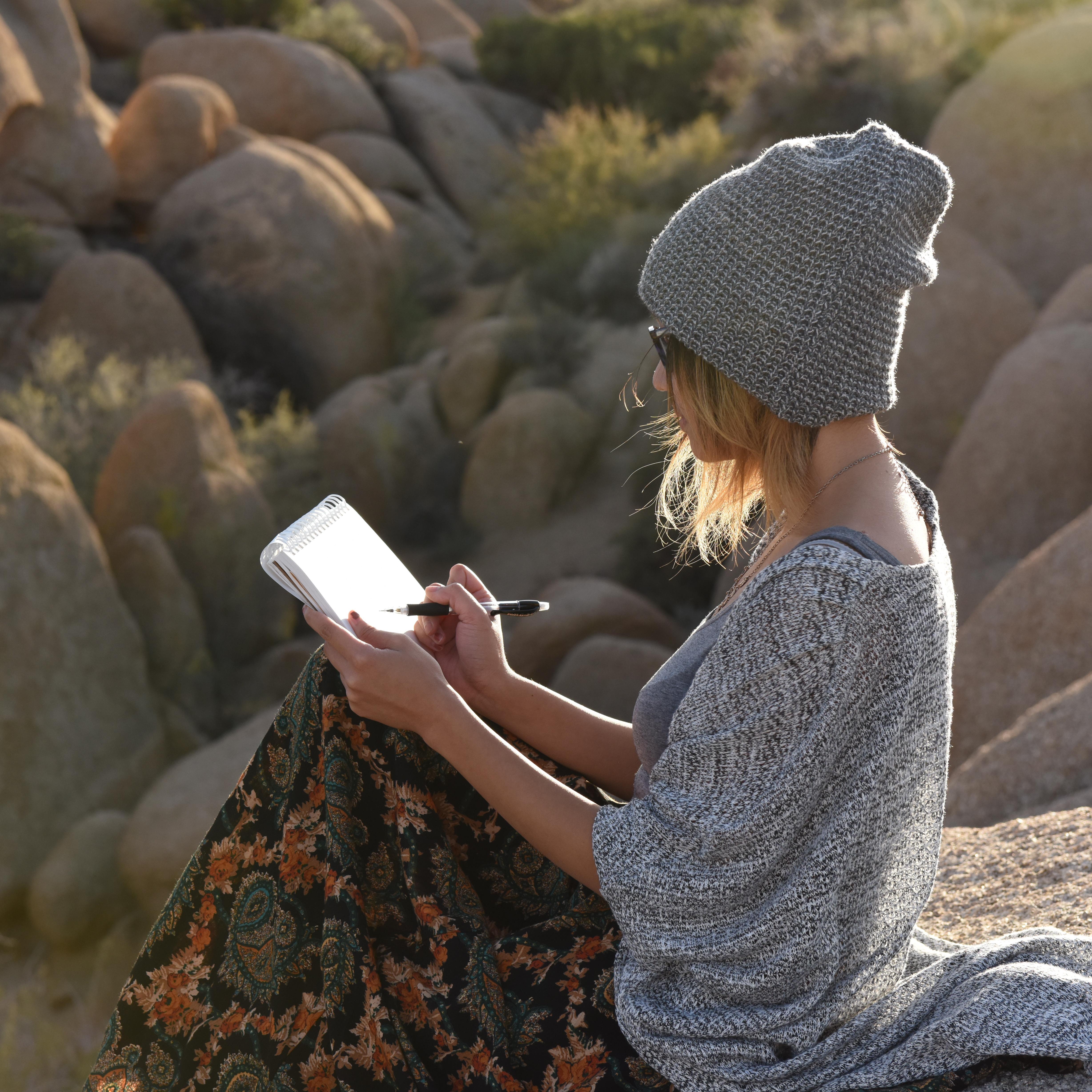 An artist sketching in a sketchpad while sitting on a large rock.