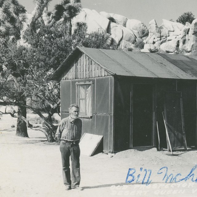 a black and white photo of a man standing in front of a cabin