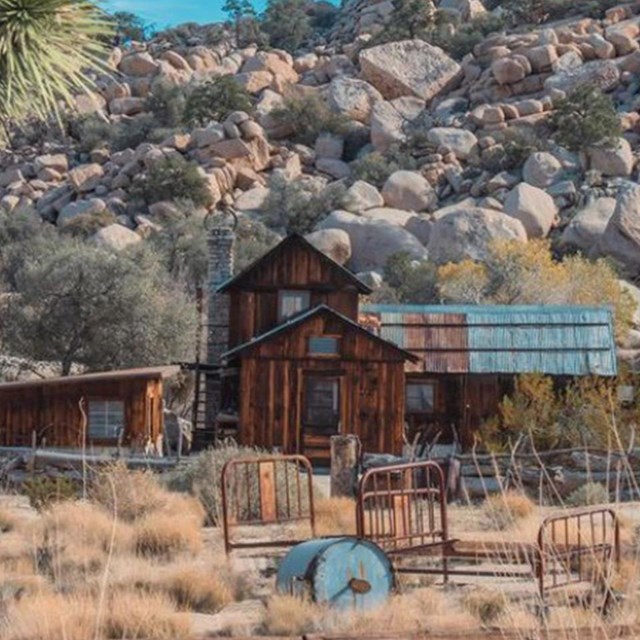 A historic wooden home with boulders and desert vegetation in the background