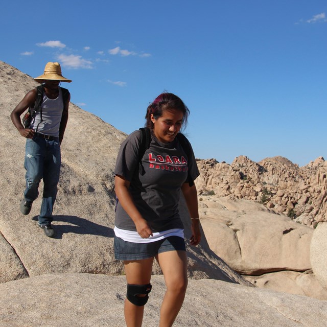 Two hikers walking on top of a rock slab with a blue sky and boulders in the background. 