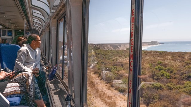 A woman sits on a train looking out of a window at the ocean below