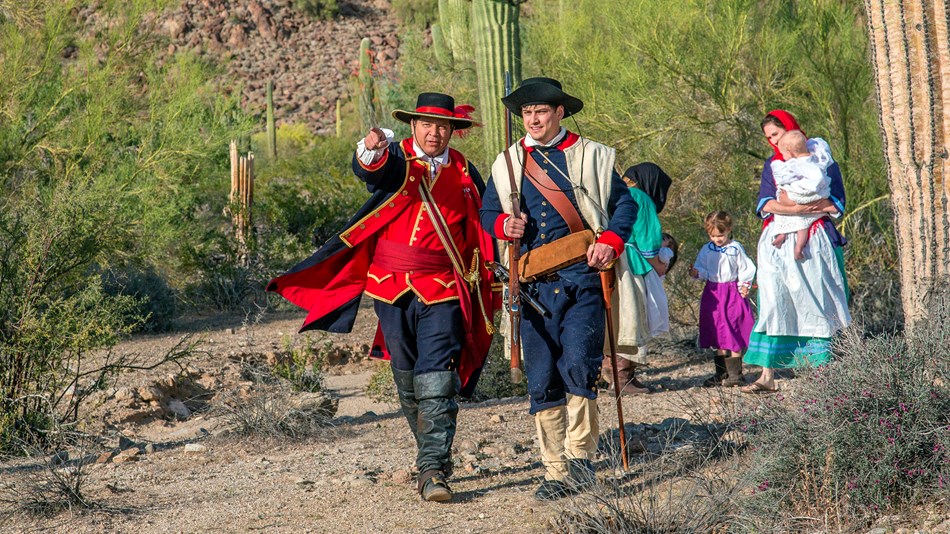 A small group of men women and children in period uniforms walk through a desert setting