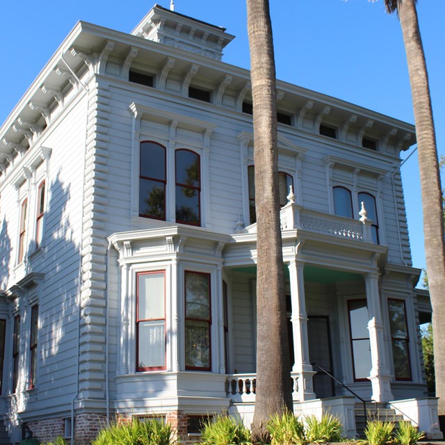 A two story Victorian home sits on a hill and is surrounded by trees. Two palm trees tower above the