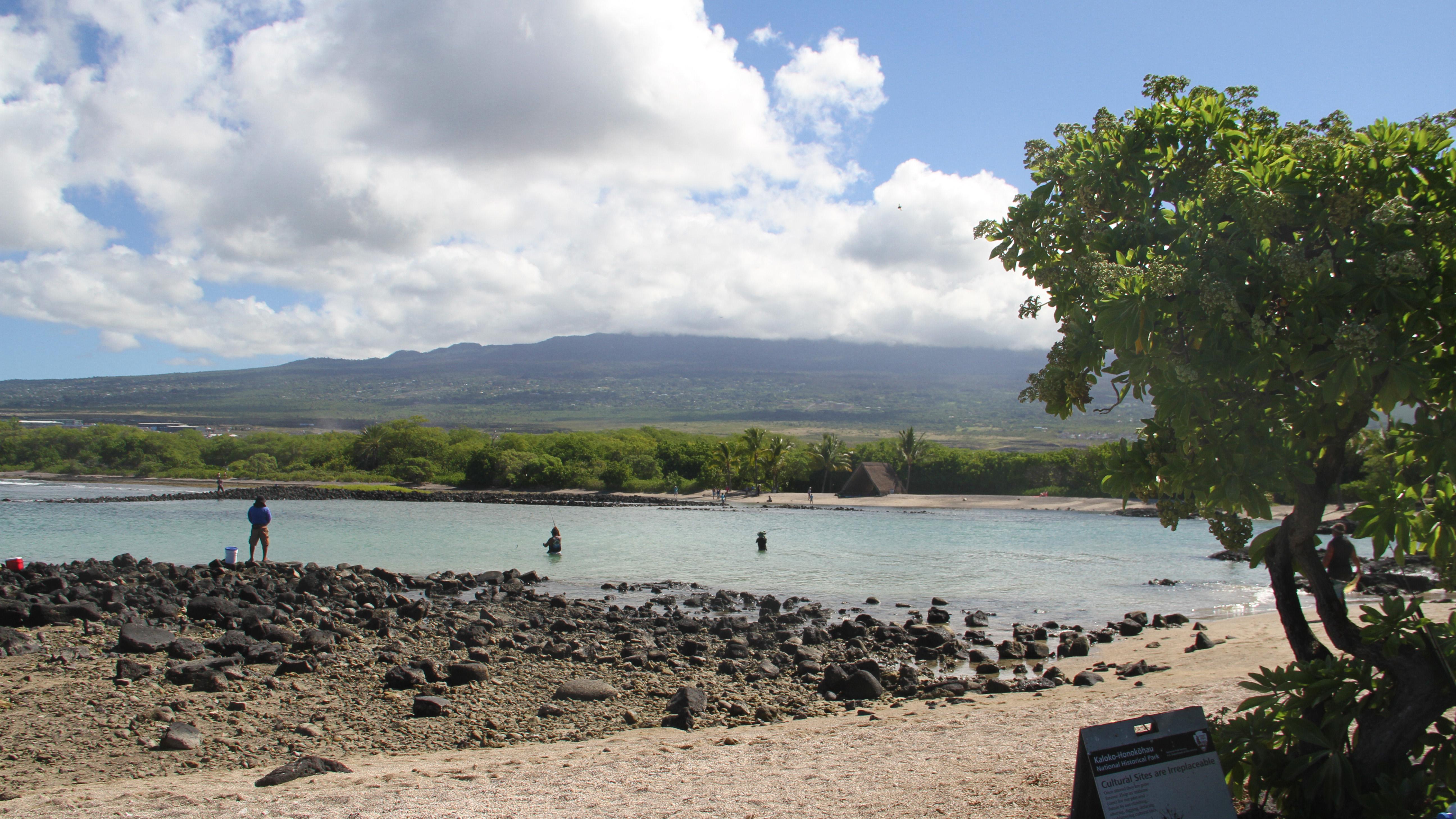 White sand and black lava in foreground, backed by a shallow bay and a distant cloud-covered volcano