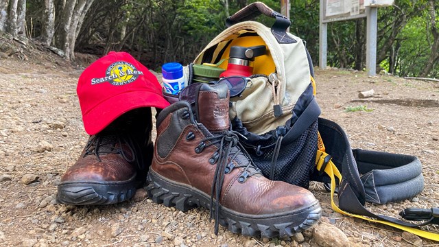 Hiking boots, a red ball cap, and a pack with water, sunglasses, and sunscreen.