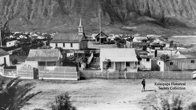 A black and white image of a town with large cliffs rising in the background. 