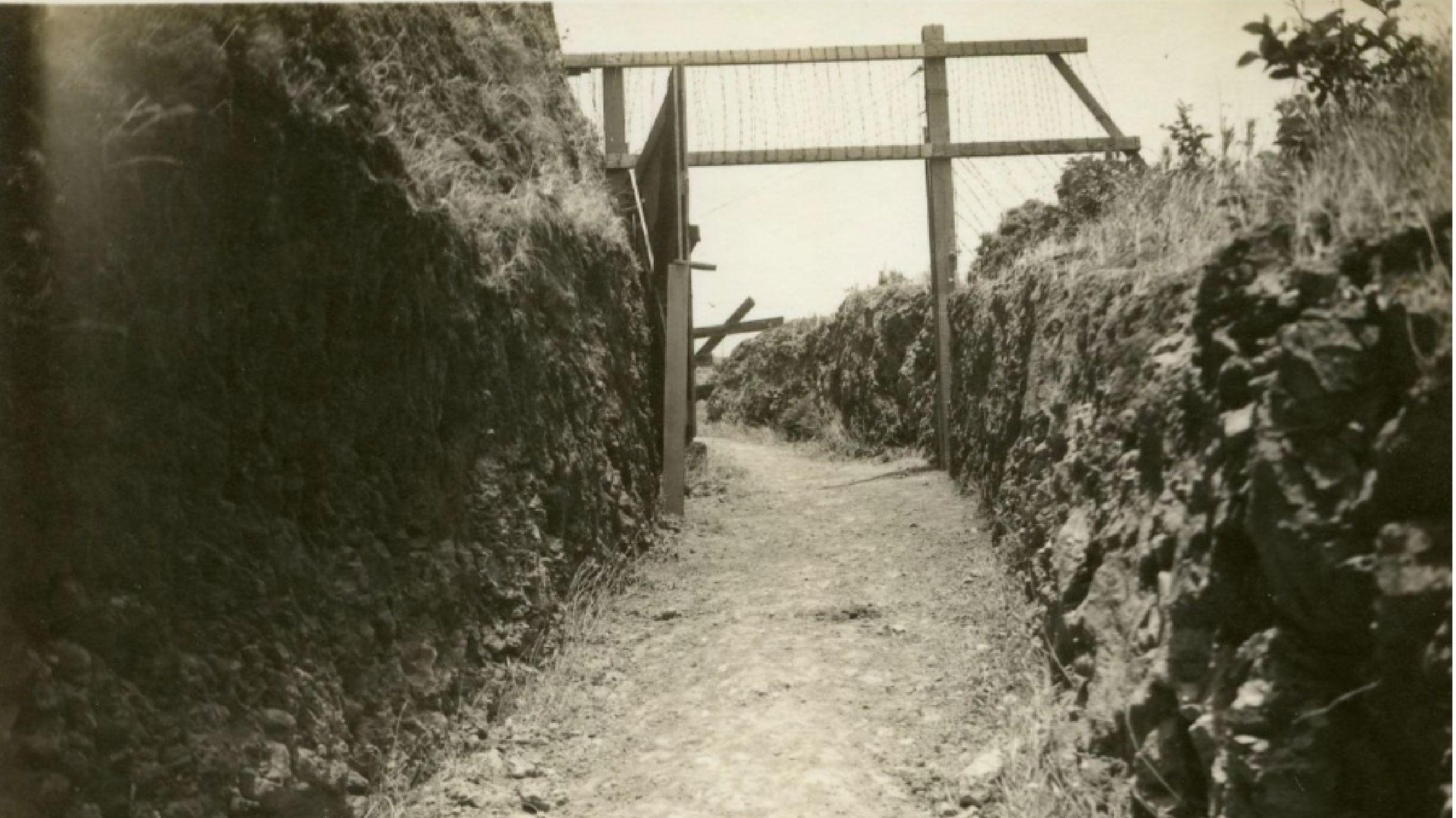 Sepia photo; outside, dirt trail leading to a gate structure with stone walls on either side.