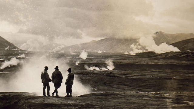 A historic photo of three men standing by a steam vent, with mountains in the background.