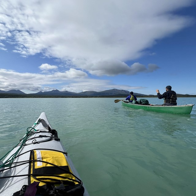the nose of a kayak on turquoise blue water. Two people paddle a canoe in the distance 