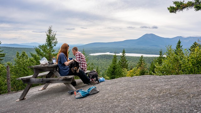 Hikers enjoy a picnic overlooking forested landscape on top of a mountain on an overcast day.