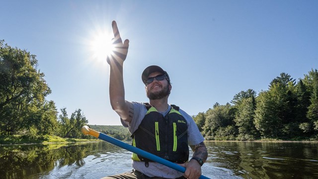 A man points something out in the distance from a canoe. The suns shines above the river behind him.