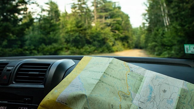 A road map sits on the dashboard of a car driving through the forest with green trees and dirt road.