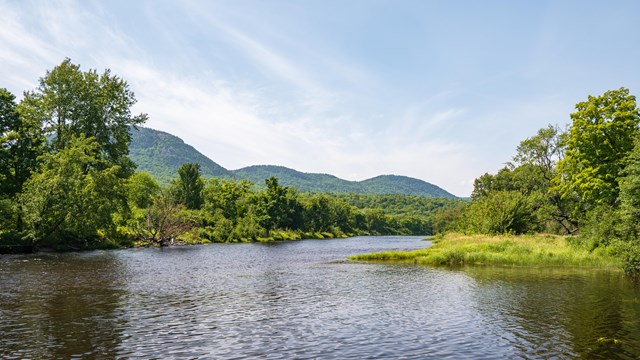 Alt text: Green mountains sit behind a flowing waterway under a blue sky. Green trees line the water