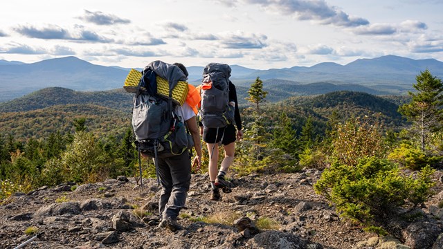Alt text: Hikers walk across rocky ground. They have backpacks and the background shows mountains.
