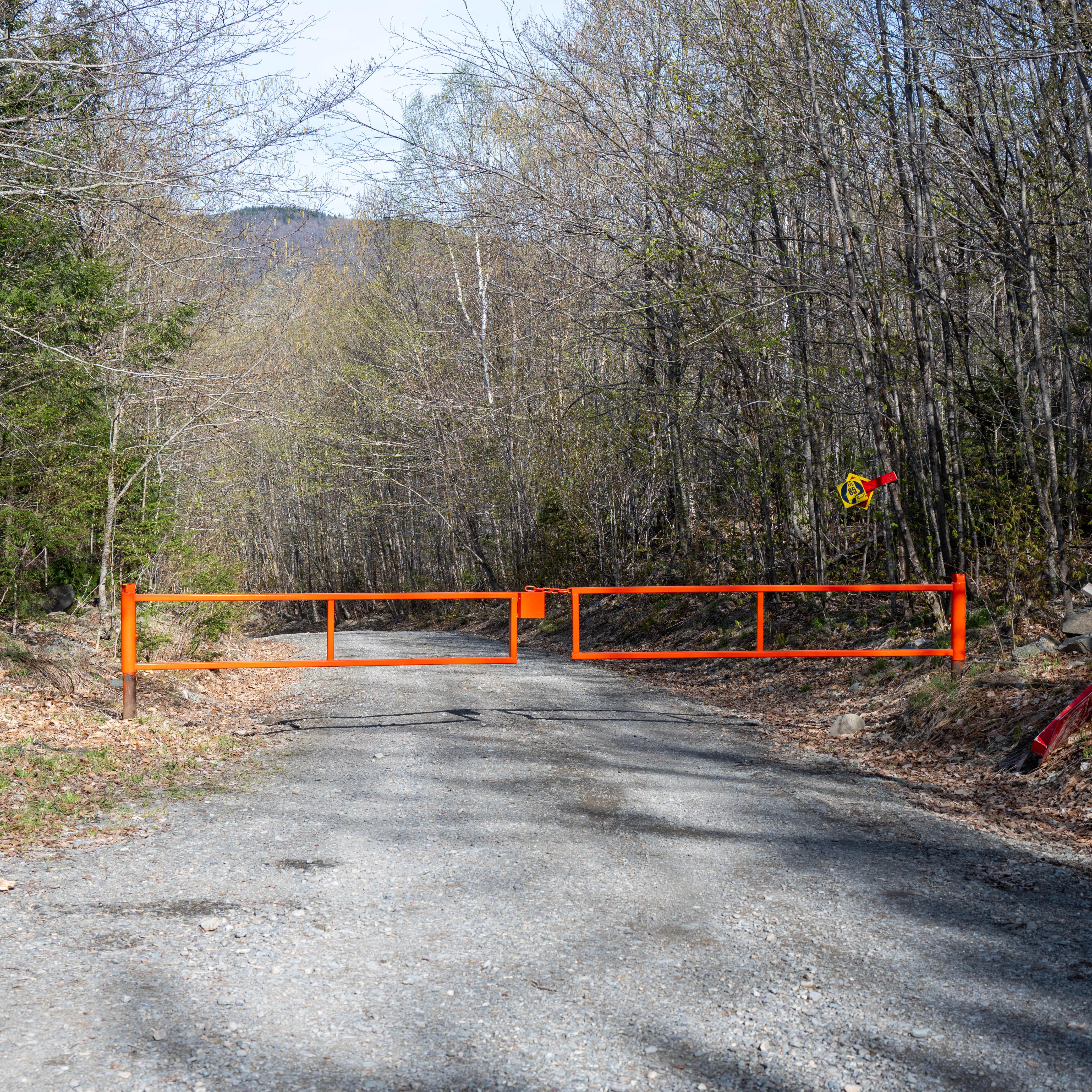 Image of a closed gate at the middle of a road. The gate is orange and the road is unpaved.