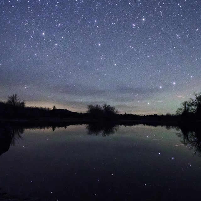 millions of stars light the night sky with the silhouettes of the woods surrounding the river in the