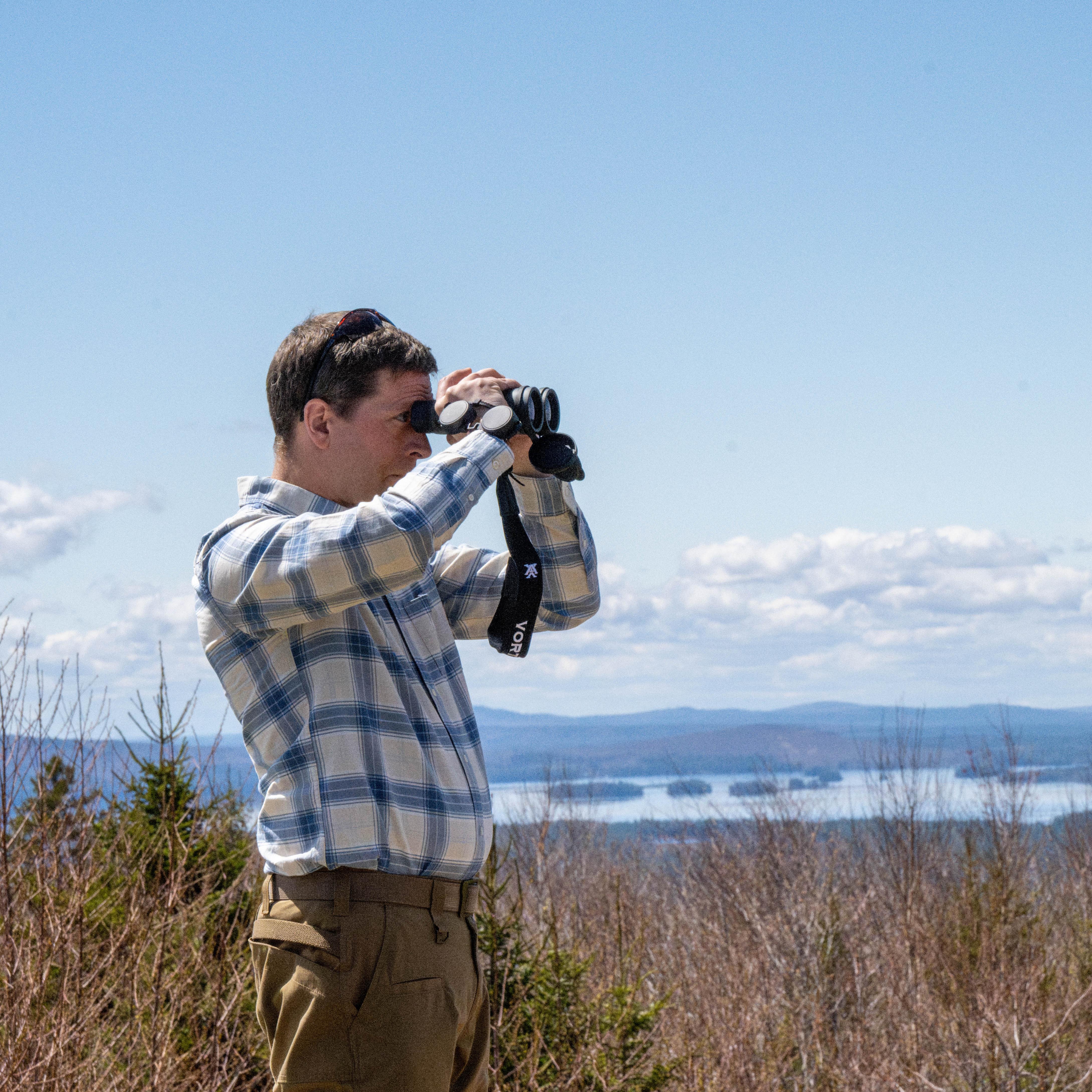 A man looks through a binocular on a sunny blue-sky day. A mountain and lake are behind him.