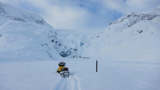 A snowmachine next to sign with glacier under snow in background