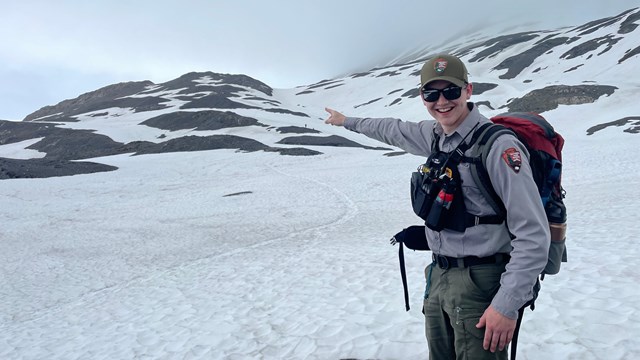 A park ranger in uniform points to snow-covered mountain terrain.