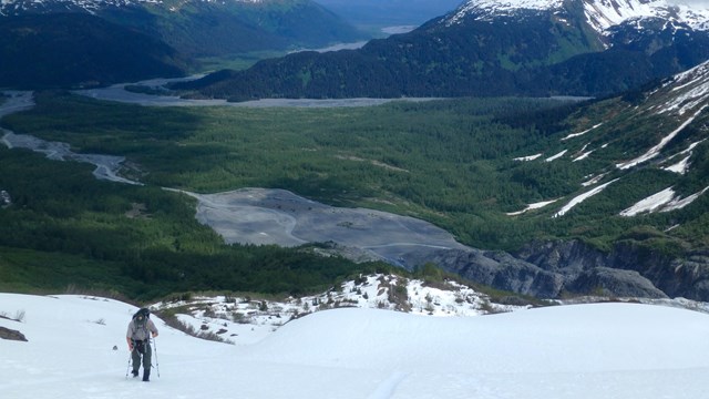 A person walks up steep snow with a green valley beyond.