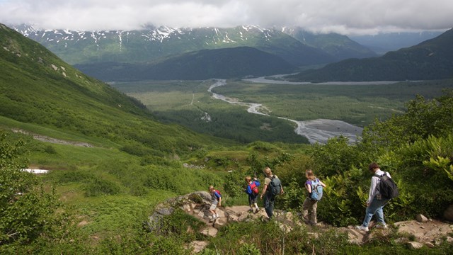 Hikers walk down a trail towards a green valley