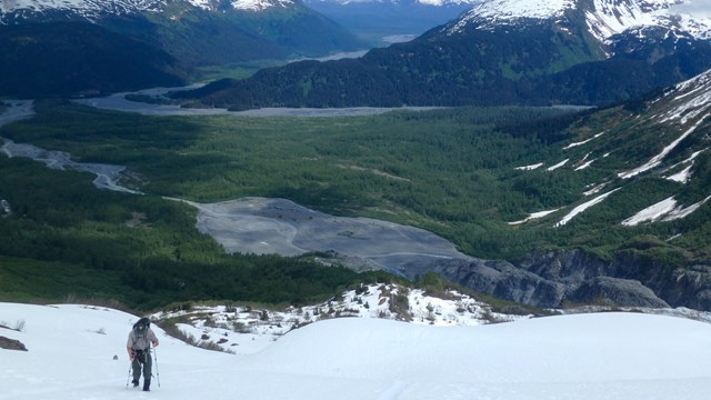 A ranger hikes up steep snow with a green valley and snow-covered mountains beyond.