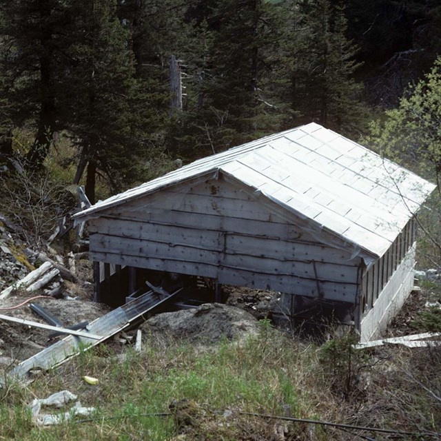 A historic wooden building with missing boards along the bottom, surrounded by woods. 