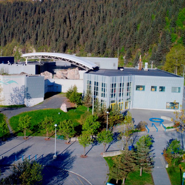 An aerial view of a grey building with the ocean to one side and green lawn and trees in the front.